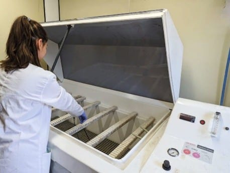 Técnica de laboratorio colocando muestras en una cámara de niebla salina para realizar un ensayo de corrosión acelerada y evaluar la resistencia de materiales.