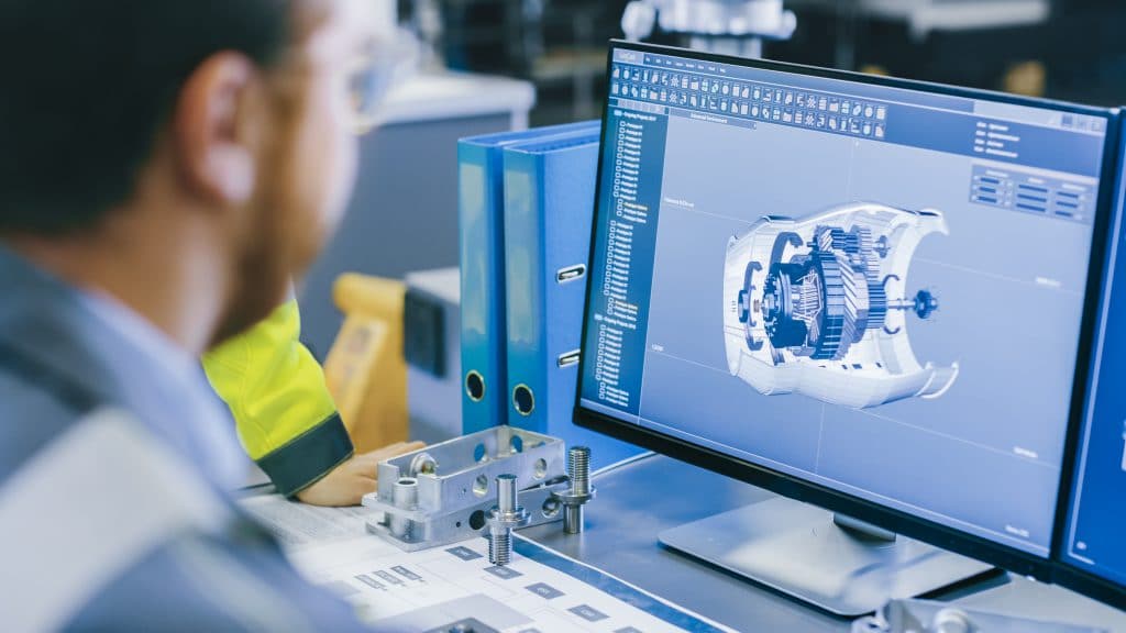 Ingeniero con ropa técnica revisando en pantalla el modelado 3D CAD de un componente mecánico industrial, con una pieza metalizada mecanizada sobre la mesa, en un entorno de desarrollo de producto.