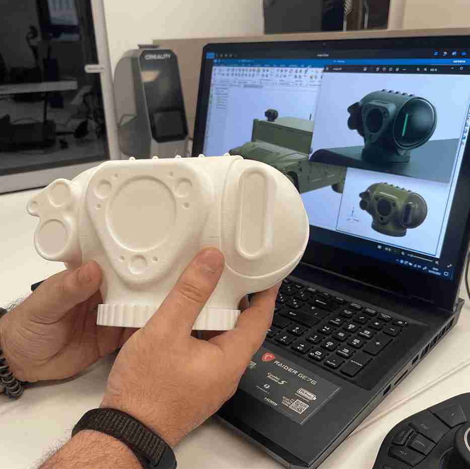 Diagnóstico inicial para desarrollo de producto Técnico sosteniendo un prototipo de carcasa impreso en 3D en material blanco, comparándolo con su modelado CAD y su render en la pantalla de un portátil, con una impresora 3D al fondo, durante el proceso de prototipado rápido.