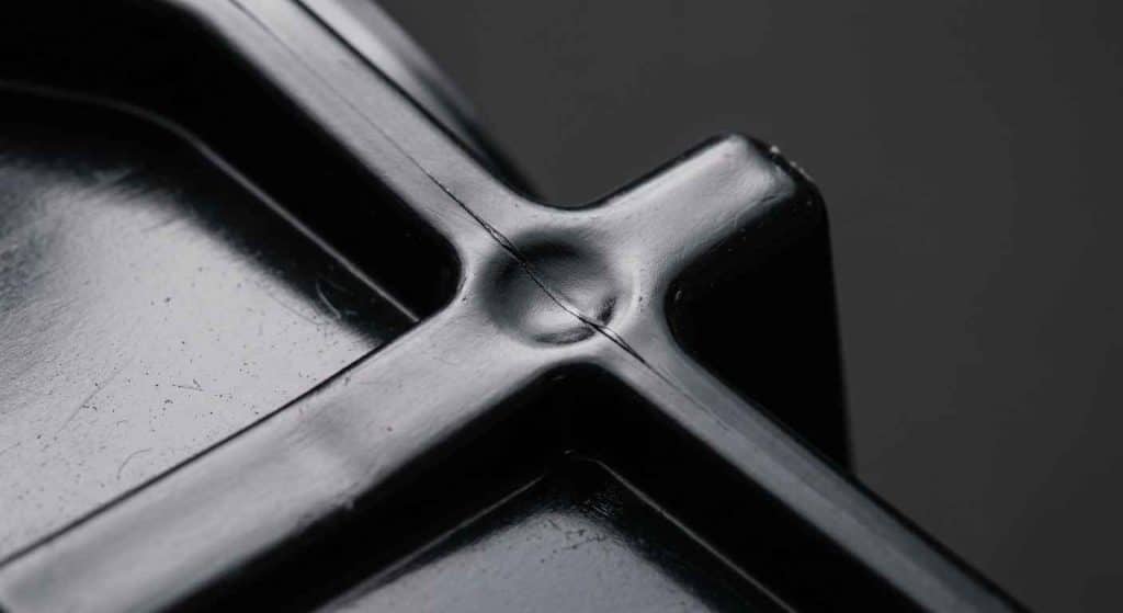 Defectos de inyección de plásticos y cómo evitar el rechazo de lote Detalle macro de una pieza inyectada en negro con una línea de soldadura y un rechupe visibles sobre una nervadura gruesa bajo luz rasante.